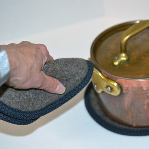 May include: A gray felt pot holder with a black trim is being held in a hand. The pot holder is being placed on top of a black pot holder that is under a copper pot with a brass handle.