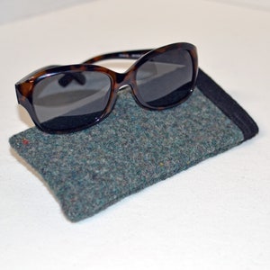 May include: Tortoiseshell sunglasses with dark lenses rest on a gray felt glasses case. The case has a black trim and a textured appearance. The sunglasses have a classic, rounded shape, suitable for eye protection.