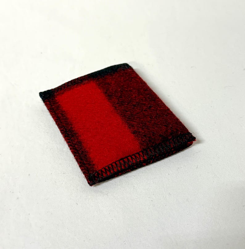 May include: A small, rectangular red and black plaid fabric item. The item has a solid red center with black plaid edges and black stitching around the perimeter. The item is likely a small pouch or case.