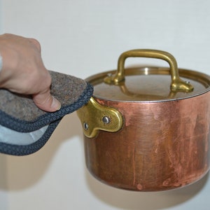 May include: A copper pot with a brass handle and lid. The pot is being held by a hand wearing a watch and a gray and black pot holder.