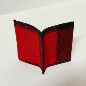 May include: A small, open book-shaped item with a red and black plaid pattern. The edges are finished with black stitching. The item is made of a soft, felt-like material.
