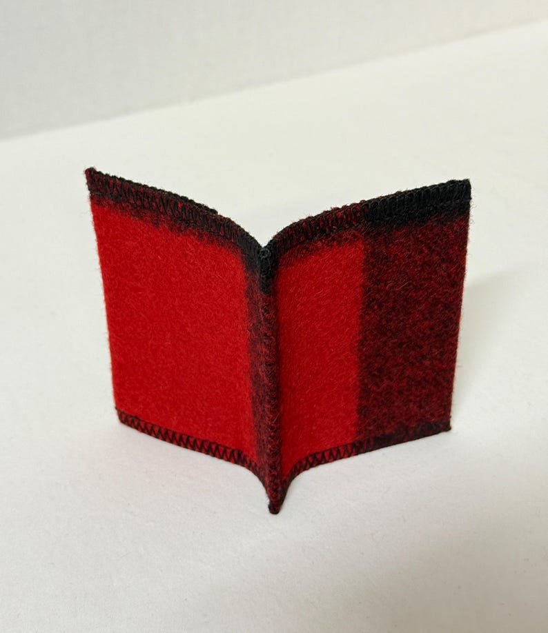 May include: A small, open book-shaped item with a red and black plaid pattern. The edges are finished with black stitching. The item is made of a soft, felt-like material.