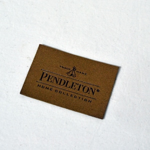 May include: A brown leather label with the text "Pendleton® Home Collection" and a logo that says "Trade Mark" with a stylized teepee design.