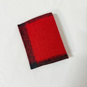 May include: A rectangular, red felt item with a black stitched border. The edges have a black and red plaid pattern. The item is likely a small wallet or card holder.