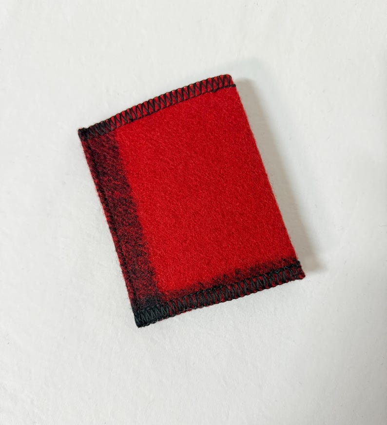 May include: A rectangular, red felt item with a black stitched border. The edges have a black and red plaid pattern. The item is likely a small wallet or card holder.