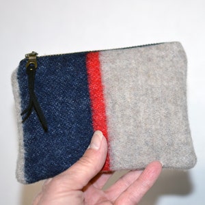 May include: A small zippered pouch made from a gray, blue, and red striped wool fabric. The pouch has a black leather zipper pull with a fringe.