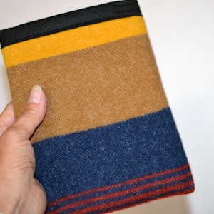 May include: A rectangular, multi-colored fabric item with horizontal stripes of black, yellow, brown, and blue. The blue section has thin red stripes. The item is held by a hand, suggesting it's a small blanket or a similar textile product.