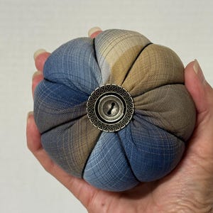 May include: A round, stuffed fabric pincushion featuring a segmented design with alternating blue and tan plaid fabric panels. The center is accented with a decorative, circular, antique-style metal button with an intricate embossed border.