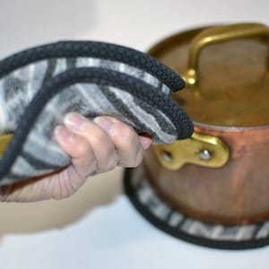 May include: A black and white striped pot holder with a black border. The pot holder is folded in half and being held by a hand. The pot holder is being used to protect a hand from a hot copper pot with a brass handle and lid.