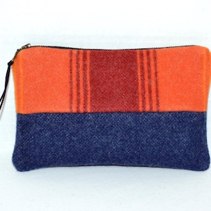 May include: A rectangular zippered pouch with a color-blocked design. The top half is orange with a central red stripe and vertical lines, while the bottom half is navy blue. A black zipper and pull complete the design.