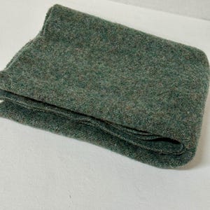 May include: A folded rectangular textile piece in a deep forest green hue. The fabric features a dense, felted, and slightly mottled texture with visible variations in green and brown fibers throughout the surface of the material.