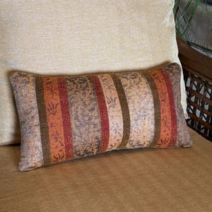 May include: Rectangular decorative pillow with a striped pattern in shades of brown, orange, and red. The pillow has a floral design in the background. The pillow is placed on a tan chair.