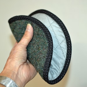 May include: A green and black pot holder with a quilted lining. The pot holder is folded in half and held in a hand.