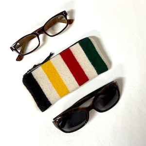 May include: A rectangular zippered pouch featuring a multi-colored striped pattern in black, yellow, cream, red, and dark green, positioned between a pair of clear-lens tortoiseshell eyeglasses and a pair of dark-lens tortoiseshell sunglasses.