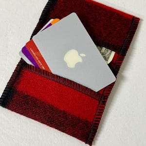 May include: Red and black plaid felt cardholder with black stitching. The cardholder contains a gray card with an apple logo, credit cards, and a dollar bill. The cardholder is rectangular and has a pocket.