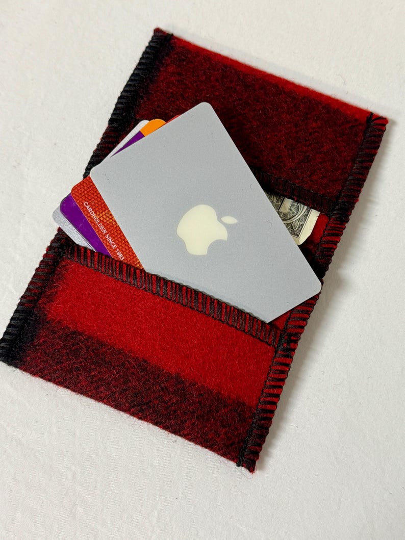 May include: Red and black plaid felt cardholder with black stitching. The cardholder contains a gray card with an apple logo, credit cards, and a dollar bill. The cardholder is rectangular and has a pocket.