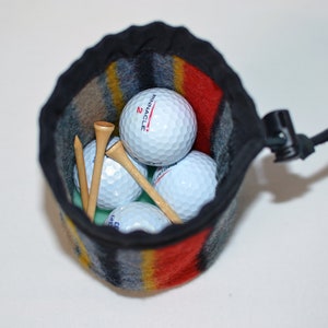 May include: A black and red striped golf ball pouch with a drawstring closure. The pouch is filled with four white golf balls and three wooden golf tees. The pouch is made of a soft, fuzzy material.