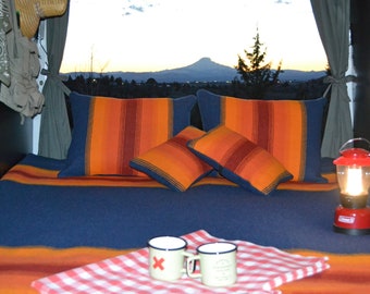 May include: A cozy camper van interior with a bed made up with a blue and orange striped blanket and pillows. The bed is facing a window with a view of a mountain range in the distance. There is a red lantern on the table next to two mugs.