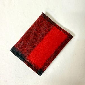 May include: A rectangular, red and black plaid fabric item. The item has a solid red stripe down the center and is bordered with black stitching. The fabric appears to be a woven material.