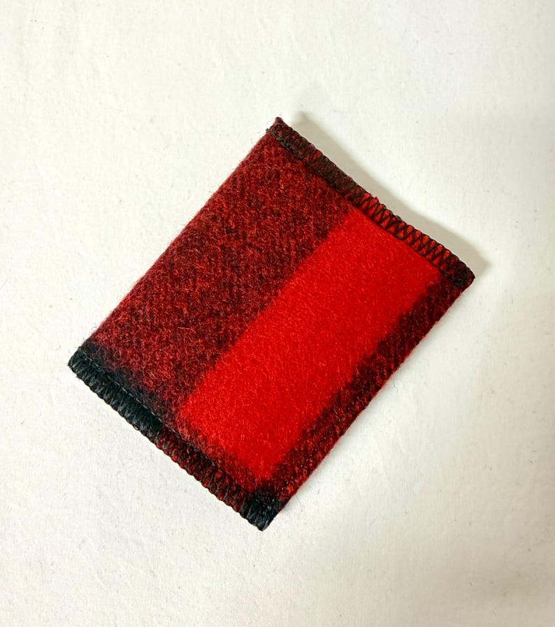 May include: A rectangular, red and black plaid fabric item. The item has a solid red stripe down the center and is bordered with black stitching. The fabric appears to be a woven material.
