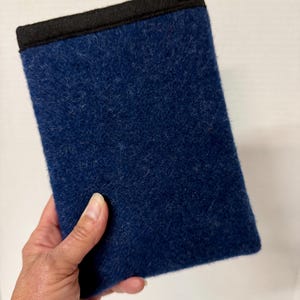 May include: A rectangular, navy blue felt case with a black trim along the top edge. The case is held in a hand against a white background. The felt has a soft, textured appearance.