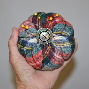 May include: A plaid fabric pincushion with a metal button in the center. The pincushion is filled with stuffing and has several pins sticking out of it.
