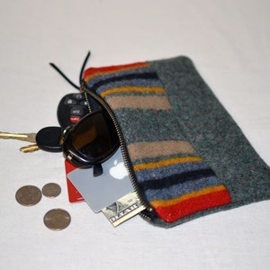 May include: A gray felt zippered pouch with a colorful striped pattern. The pouch is open and contains sunglasses, keys, a credit card, and a dollar bill.