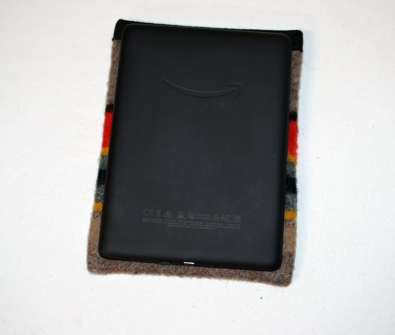 May include: A black e-reader with a gray fabric case. The case has a colorful striped pattern with red, yellow, blue, and gray stripes. The e-reader has the Amazon logo on the front.