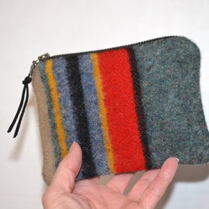 May include: A small, rectangular zippered pouch with a colorful striped design. The stripes include red, blue, yellow, and black. The pouch is made of a textured material and has a black leather pull tab on the zipper.