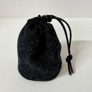 May include: A small, dark charcoal gray felt drawstring pouch with a textured, fuzzy surface. The bag features a black fabric top closure secured by a black plastic cord lock and a thin black leather cord with a knotted end for easy opening.