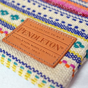 May include: A close-up of a brown leather patch with the text "WARRANTED TO BE A PENDLETON® PENDLETON WOOLEN MILLS SINCE 1863" on a white and blue striped fabric with a colorful geometric pattern.