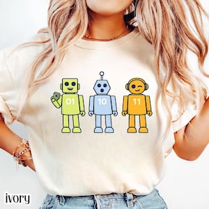 May include: Ivory-colored sweatshirt featuring three colorful robot illustrations. The robots are yellow, blue, and orange, each with a unique design and number. The text "ivory" is printed at the bottom.