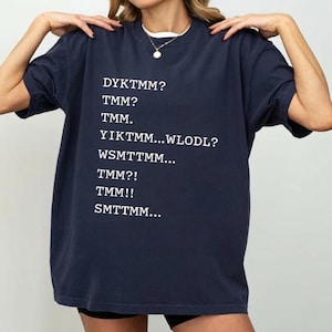 May include: Navy blue t-shirt with white text. The text includes the phrases: DYKTMM?, TMM?, TMM., YIKTMM...WLODL?, WSMTTMM..., TMM?!, TMM!!, and SMTTMM... The shirt has short sleeves and a crew neck.