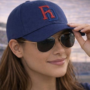 May include: A navy blue baseball cap with a red and blue embroidered logo. The cap is worn with gold-framed aviator sunglasses. The person is touching the brim of the cap.