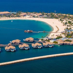 May include: Aerial view of a tropical resort with overwater bungalows and beachfront villas. The turquoise water surrounds the island, with a sandy beach and lush greenery. Brown rooftops and white buildings are visible.