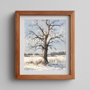 Puede incluir: Pintura al óleo de un árbol desnudo en un campo nevado, enmarcada en un marco de madera marrón claro. La obra representa un paisaje invernal con un cielo azul y un campo cubierto de nieve. El árbol es el punto focal.