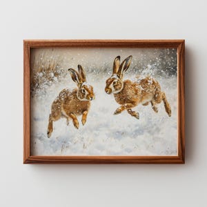 Puede incluir: Una obra de arte enmarcada representa dos liebres marrones corriendo por un paisaje nevado. Las liebres están en plena carrera, con nieve en su pelaje. La obra está dentro de un marco de madera marrón.