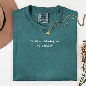 May include: Teal Comfort Colors t-shirt with the text "return this.degree to society" in white. A gold necklace with a star pendant rests on the shirt. A brown hat and sprigs of pink flowers are also in the image.