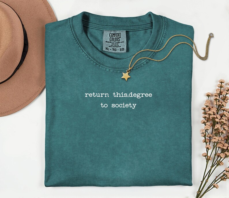 May include: Teal Comfort Colors t-shirt with the text "return this.degree to society" in white. A gold necklace with a star pendant rests on the shirt. A brown hat and sprigs of pink flowers are also in the image.