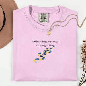May include: A light pink t-shirt with the text "Python-ing my way through life" and a graphic of a blue and yellow snake. A gold necklace with a star pendant is draped on the shirt.