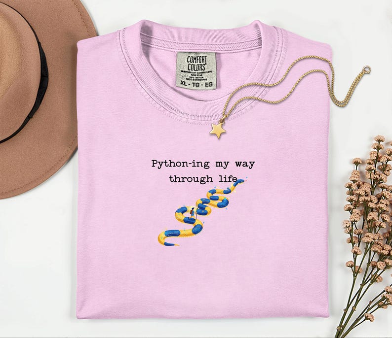 May include: A light pink t-shirt with the text "Python-ing my way through life" and a graphic of a blue and yellow snake. A gold necklace with a star pendant is draped on the shirt.