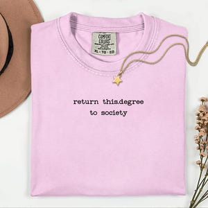 May include: A light pink t-shirt with the text "return this.degree to society" in black. A gold-colored necklace with a star pendant rests on the shirt. A brown hat is visible in the upper left corner.