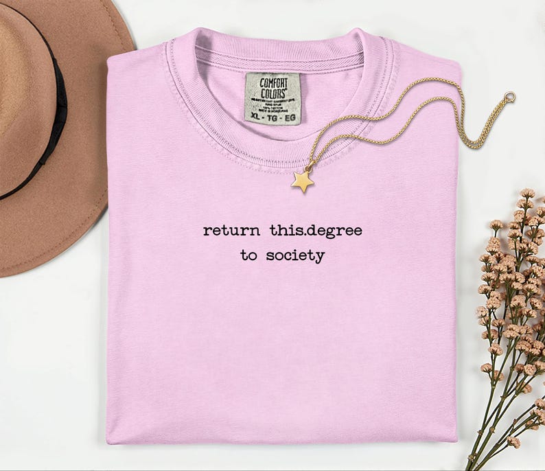 May include: A light pink t-shirt with the text "return this.degree to society" in black. A gold-colored necklace with a star pendant rests on the shirt. A brown hat is visible in the upper left corner.