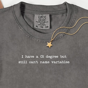 Peut inclure: T-shirt gris Comfort Colors avec le texte "I have a CS degree but still can't name variables." Un pendentif étoile doré est suspendu à une chaîne délicate. Le t-shirt a un col rond et une étiquette XL-TG-EG.