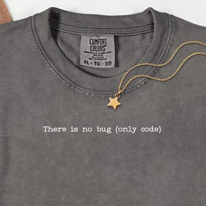 May include: Charcoal grey t-shirt featuring the phrase "There is no bug (only code)". The shirt has a crew neck and a label reading "Comfort Colors". A gold star pendant hangs from a fine chain.