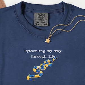 May include: Navy blue t-shirt with the text "Python-ing my way through life" and a graphic of a yellow and blue snake. A gold star pendant hangs from a gold chain. The shirt has a Comfort Colors tag.