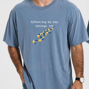 May include: Blue t-shirt with the text "Python-ing my way through life" above a graphic of yellow and blue inflatable tubes. The shirt is a solid blue color and has short sleeves.
