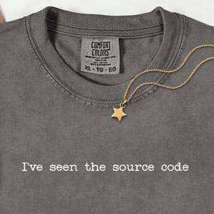 Könnte beinhalten: Ein graues Comfort Colors T-Shirt mit dem weißen Text "I've seen the source code". Ein goldener Sternanhänger hängt an einer zarten Kette. Das Shirt hat einen Rundhalsausschnitt und ein Etikett mit der Aufschrift XL-TG-EG.