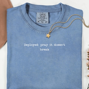 May include: A light blue t-shirt with the text "Deployed: pray it doesn't break" in white. A gold-colored star pendant necklace rests on the shirt. The shirt has a Comfort Colors tag.