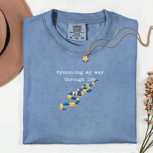 May include: A light blue Comfort Colors t-shirt with the text "Python-ing my way through life" and a yellow and blue snake graphic. A gold star pendant necklace rests on the shirt. A brown hat and flowers are in the background.